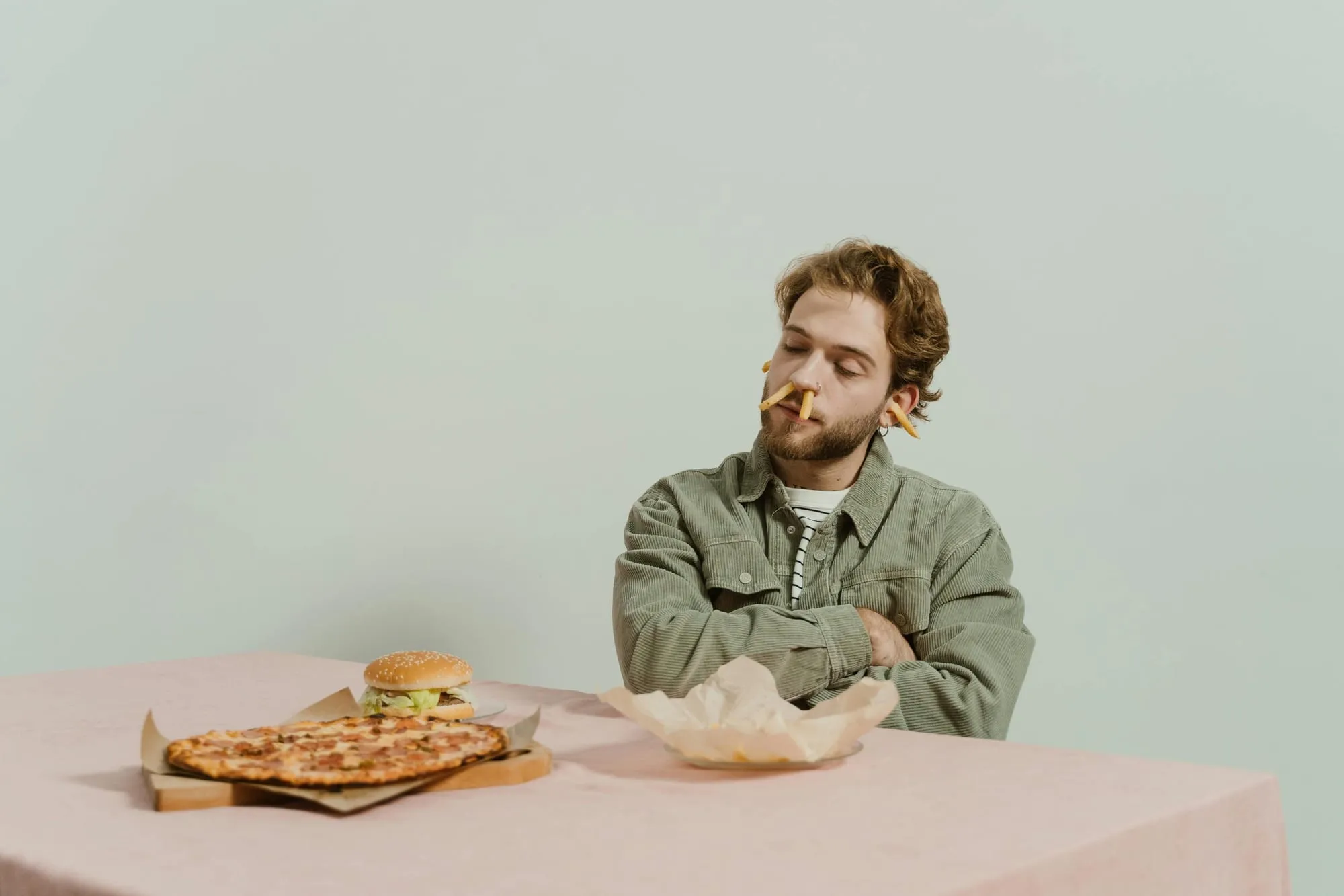 A young man at a table declining pizza and a burger, with french fries protruding from his nose and ears